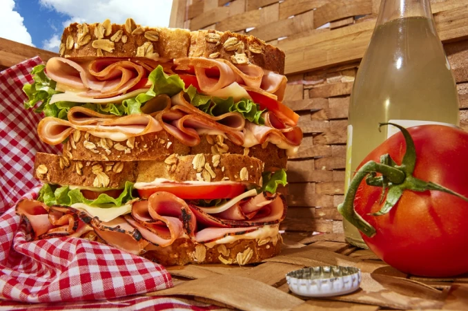 Towering multi-layer sandwich with sliced turkey, tomato, lettuce, cheese, and whole-grain bread next to a soda bottle and tomato.