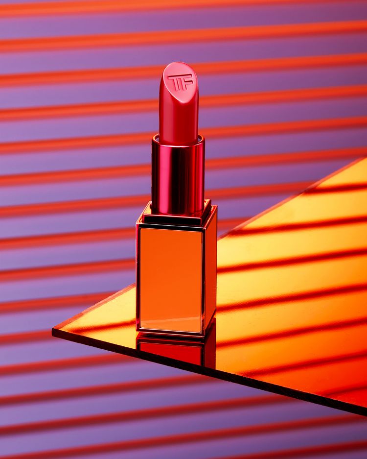 Close-up of a red lipstick tube with gold accents on a reflective surface against a striped background; beauty and makeup.