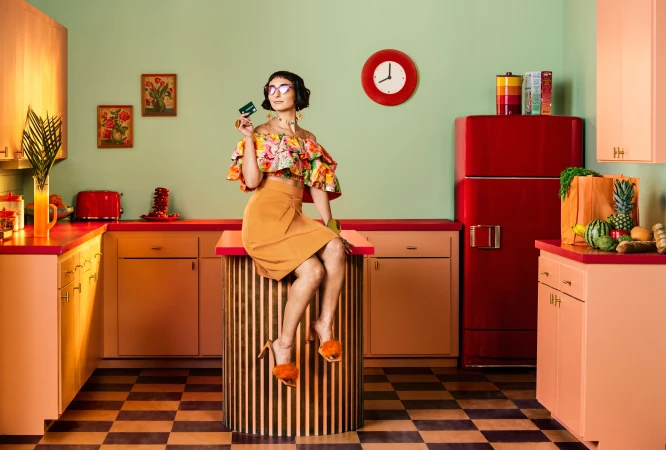 Stylish woman in floral top and orange skirt sits in retro kitchen with a credit card, showcasing vibrant colors and vintage decor.