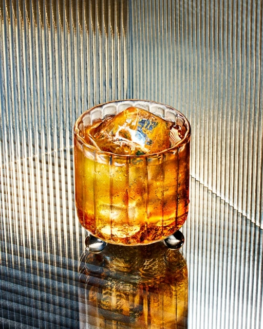 A glass filled with a golden liquid and ice sits on a reflective surface, with a textured, vertically striped background. Food & Beverage.