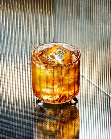 A glass filled with a golden liquid and ice sits on a reflective surface, with a textured, vertically striped background. Food & Beverage.