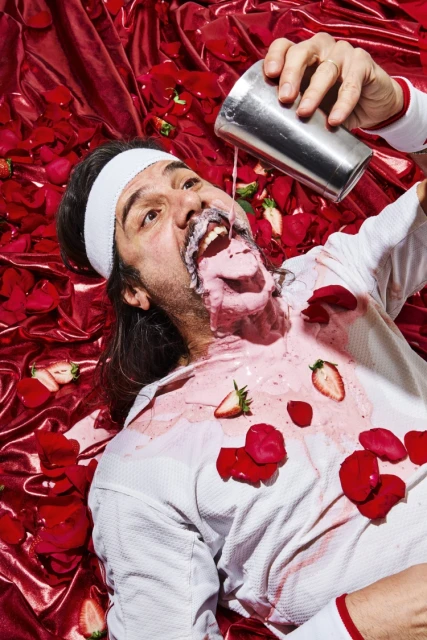 An adult male lies on a red fabric background covered in red rose petals, a beverage from a metal cocktail shaker spilling over him.