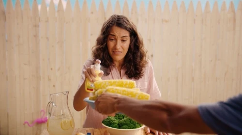 Woman spraying cooking oil on corn on the cob at an outdoor picnic table with lemonade and broccoli, set against a wooden fence.