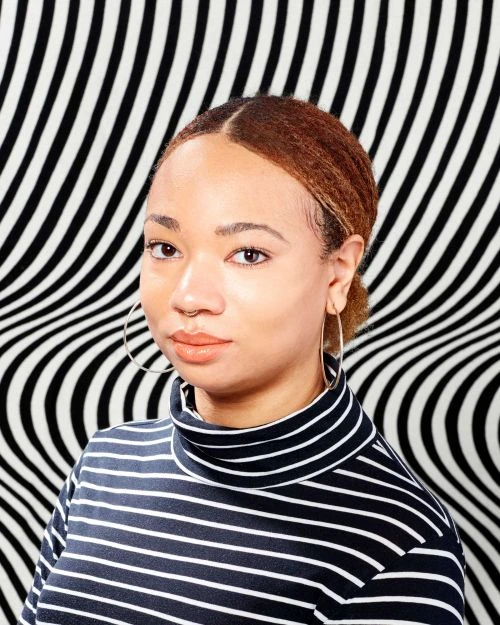 Portrait of an adult female with fair skin wearing a striped turtleneck against an abstract black and white patterned backdrop.