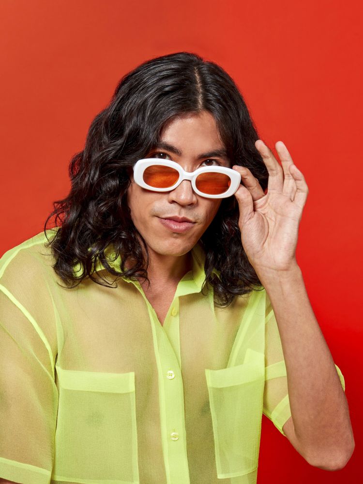 Portrait of a young adult with long black hair wearing a sheer yellow shirt and white-rimmed sunglasses against a red background.