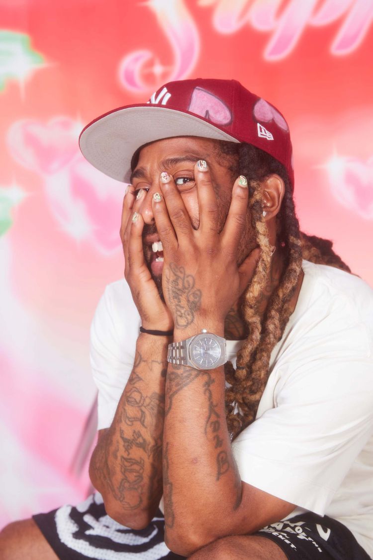 Man wearing burgundy baseball cap and white t-shirt holding hands to face against a pink airbrushed background