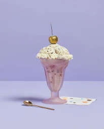An ice cream sundae in a pink glass topped with a gold cherry, placed next to a gold spoon on a light purple surface.