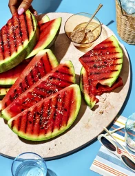 Top-down view of grilled watermelon slices on a round tray with a small bowl of seasoning and sunglasses on a blue background