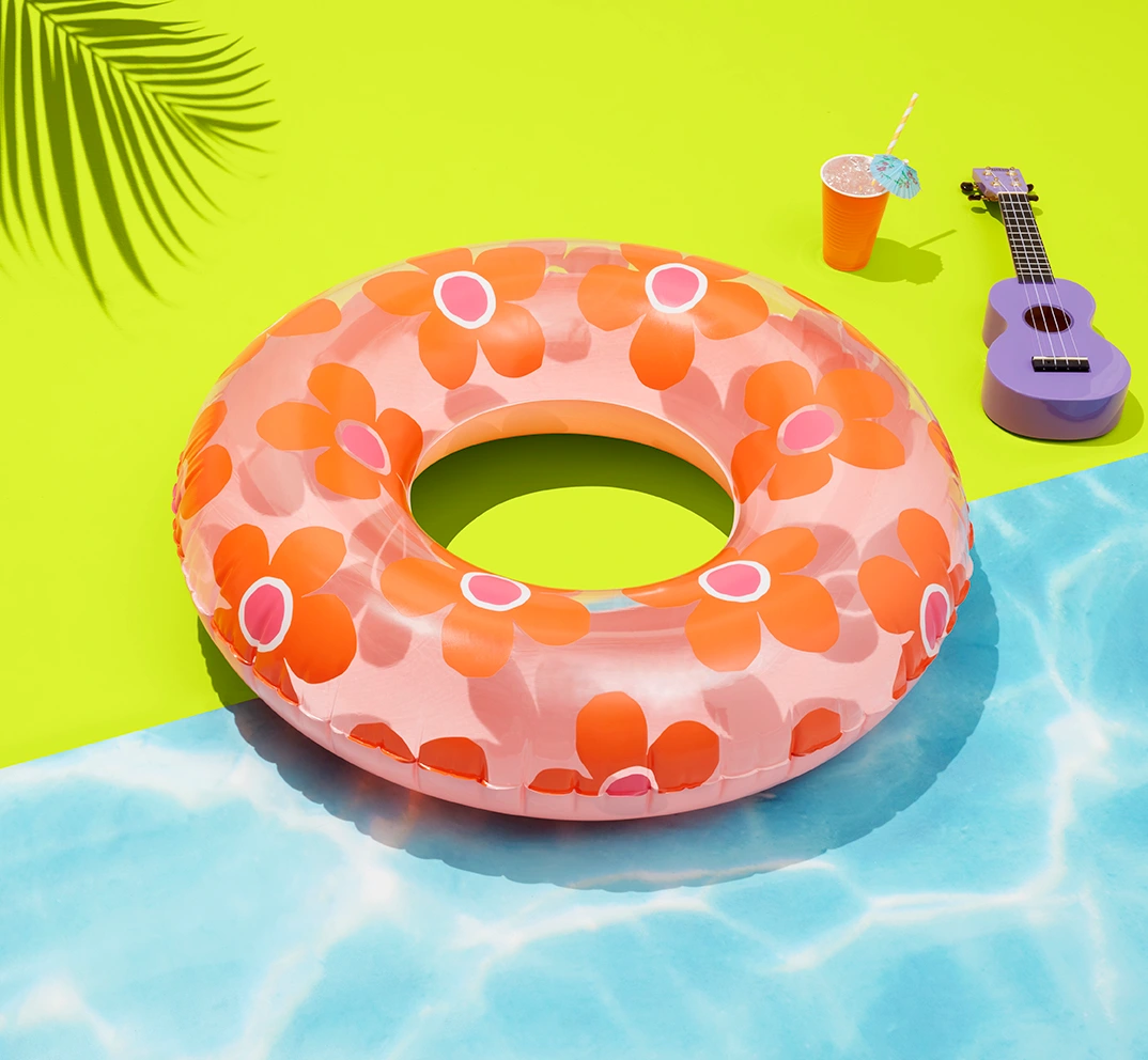 Pink and orange floral print inflatable inner tube pool float on a bright yellow and blue surface with a ukulele and drink