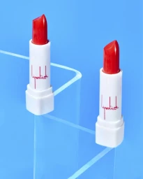 Two red lipsticks in white tubes with script text displayed on clear acrylic risers against a bright blue background