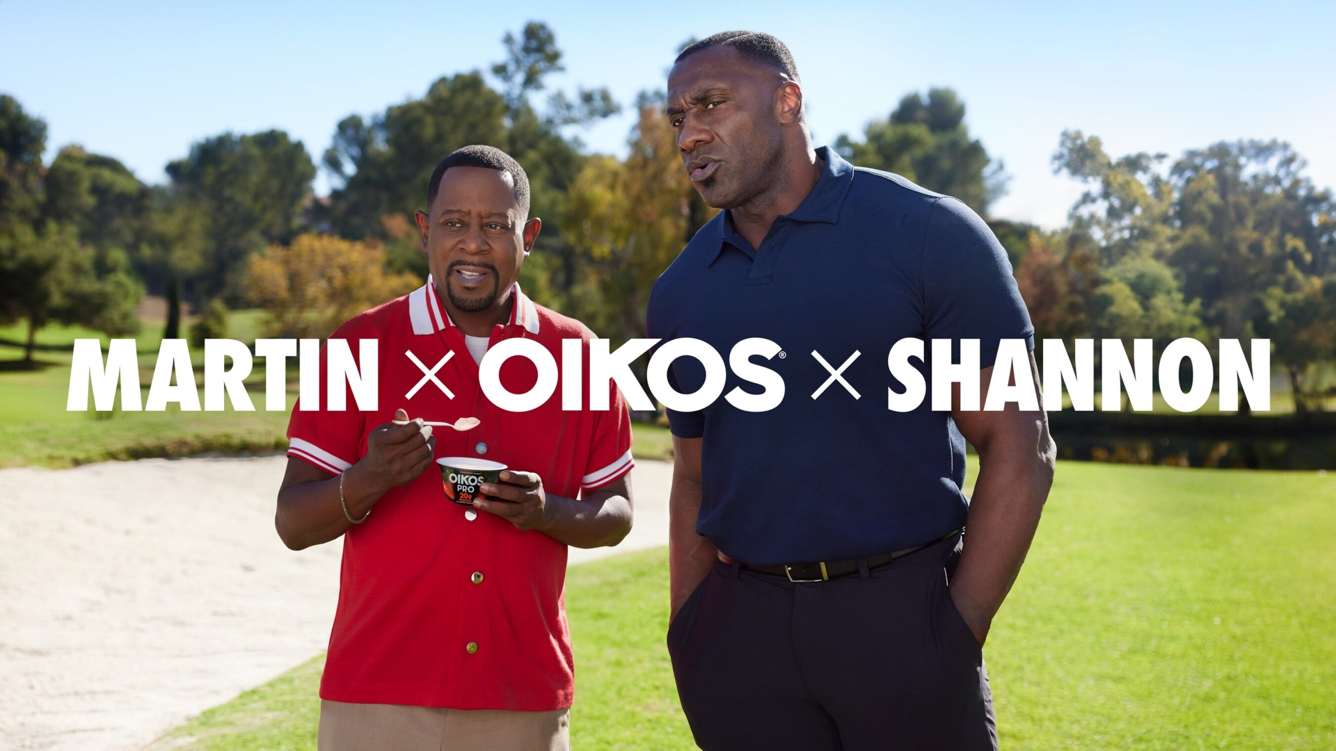 Two adult men, one in red and one in navy, are standing outdoors on a golf course with trees and a blue sky visible in the background.