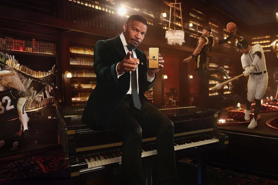 Adult male in a suit seated at a piano points while holding a gold-colored cell phone in an elegant room with sports figures.
