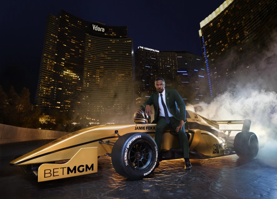 Jamie Foxx sits on a gold race car in front of the Vdara and Cosmopolitan hotels in Las Vegas at night with dramatic lighting.