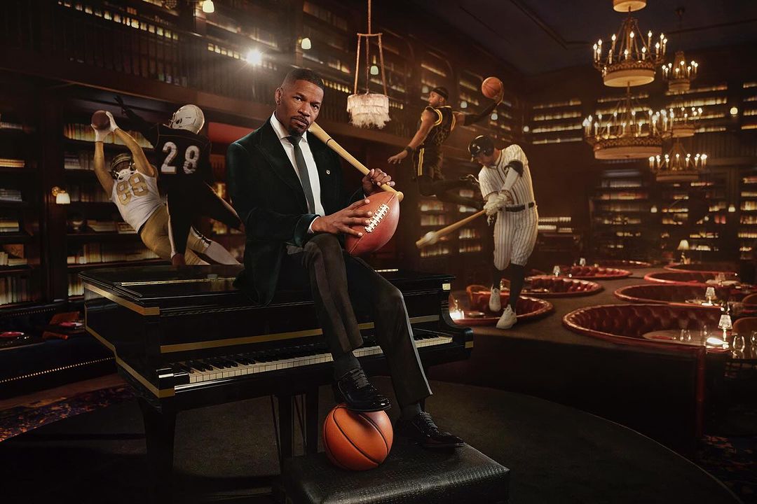 Man in a green velvet suit seated on a piano with sports imagery behind him, including baseball, football and basketball players.