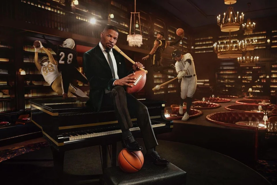 Man in a green velvet suit seated on a piano with sports imagery behind him, including baseball, football and basketball players.