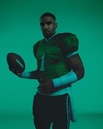 Portrait of a football player in uniform holding a football, standing against a solid green background, studio lighting.