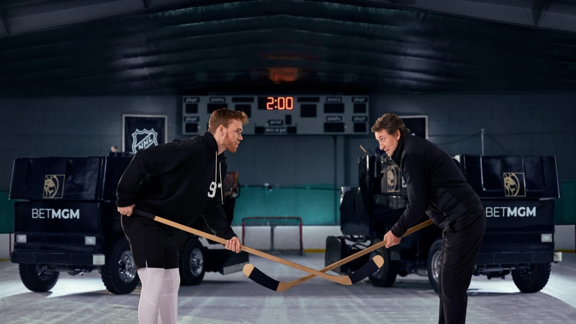 Two male hockey players face off on ice with hockey sticks between branded vehicles in a dim indoor hockey arena, a digital scoreboard reads 2:00.