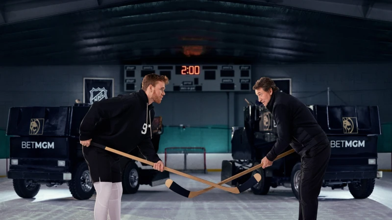 Two male hockey players face off on ice with hockey sticks between branded vehicles in a dim indoor hockey arena, a digital scoreboard reads 2:00.