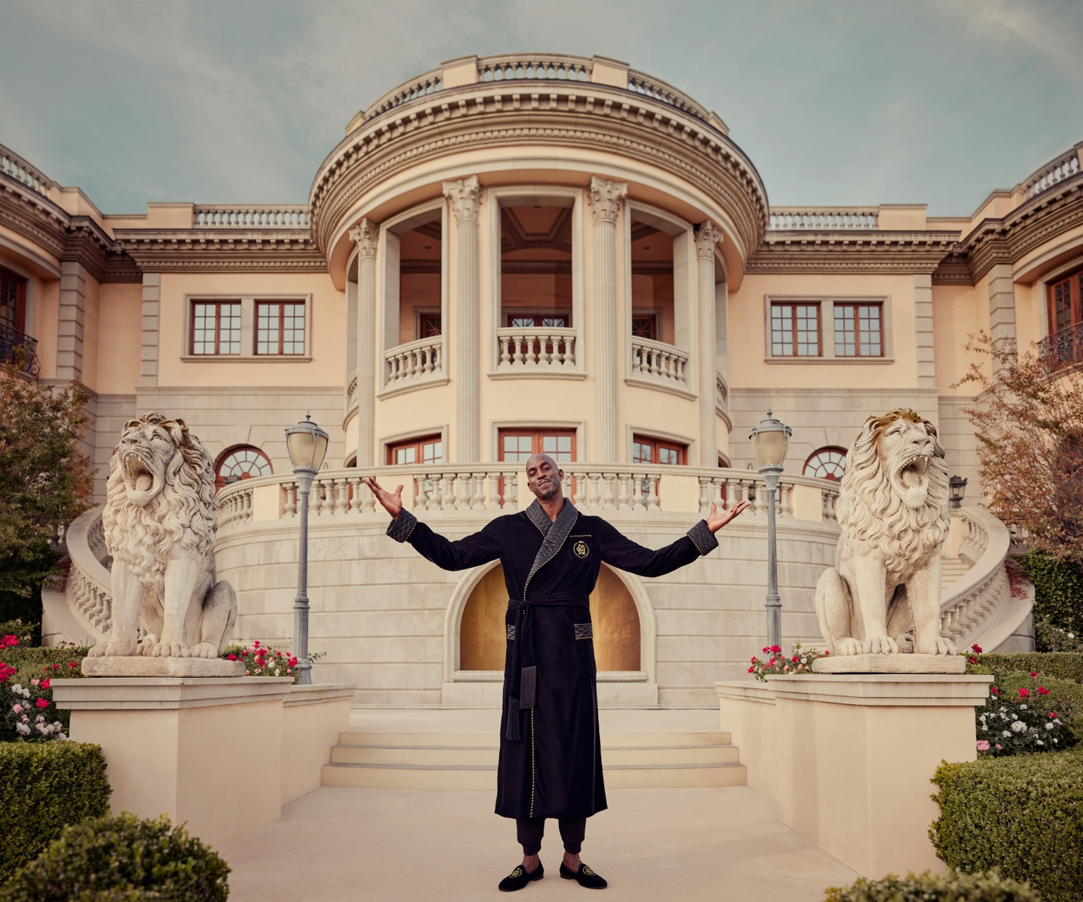 A dark-skinned man in a black robe stands in front of a large, ornate mansion with lion statues flanking the entrance staircase and garden.