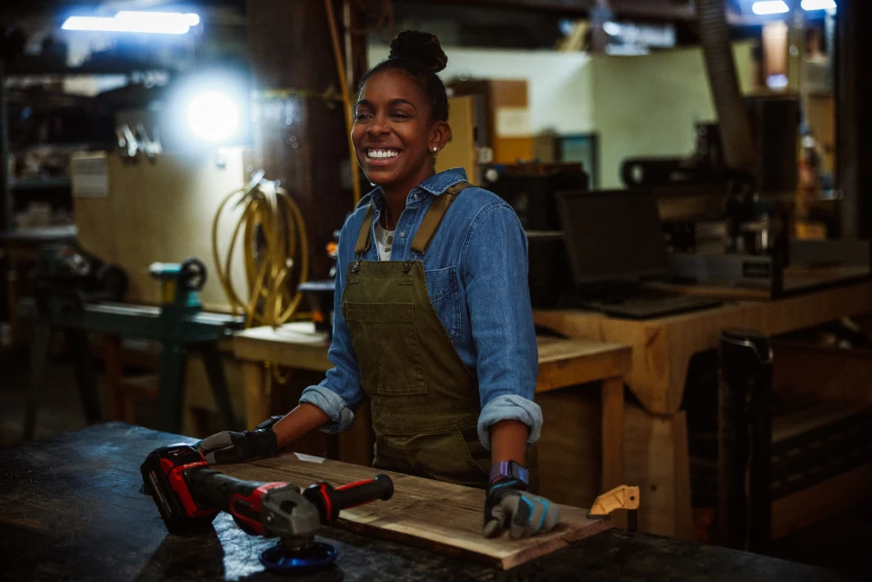 Smiling woman in a woodworking shop wearing overalls and gloves, standing at a workbench with power tools and a piece of wood.