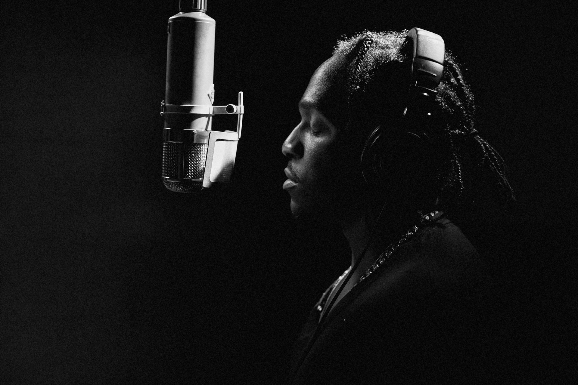 Monochrome portrait of a singer wearing headphones in a dark recording studio setting, standing next to a microphone, and singing.