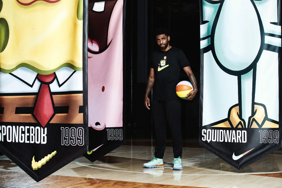 Adult male posing with a basketball in front of three cartoon character banners on a basketball court wearing a black t-shirt.