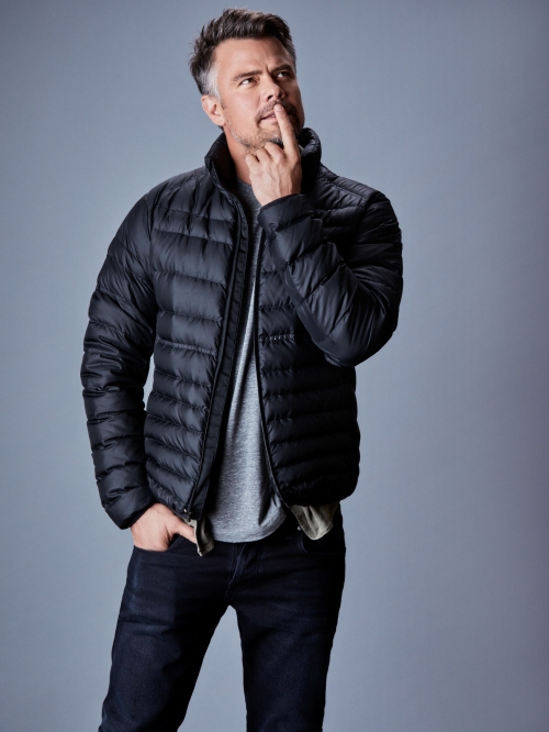 Man in a black puffer jacket stands indoors with hand on chin, wearing a gray tee shirt and jeans, looking thoughtful against a gray background.