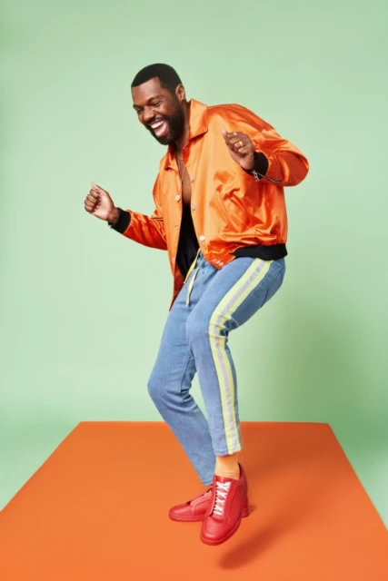 Full-length shot of an adult man with dark hair laughing while dancing on an orange platform in an orange jacket and light blue jeans.