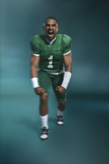 An adult male in green football uniform, with number '1', shouts with clenched fists against a blurred teal background.