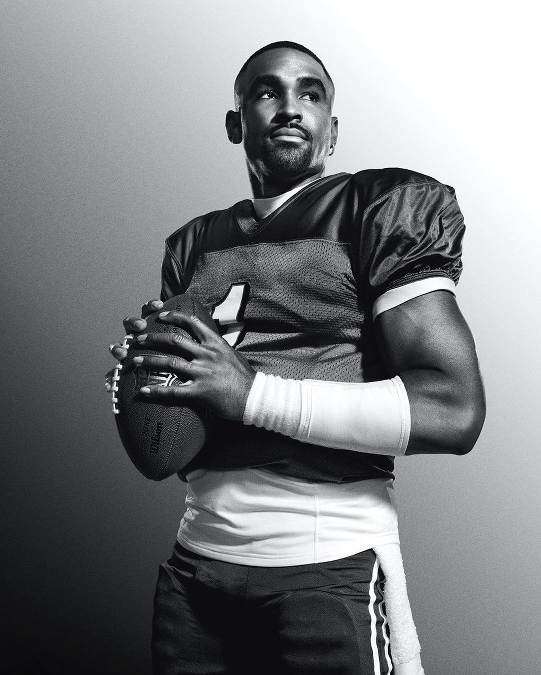 Black and white portrait of a football player in uniform, holding a football, looking upward with a concentrated facial expression.
