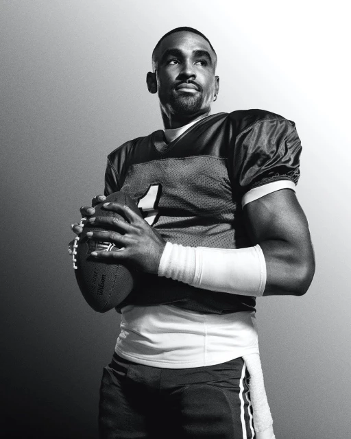 Black and white portrait of a football player in uniform, holding a football, looking upward with a concentrated facial expression.