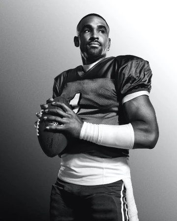 Black and white portrait of a football player in uniform, holding a football, looking upward with a concentrated facial expression.
