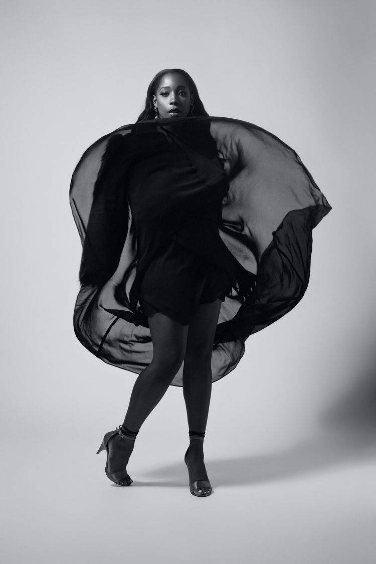 A Black woman in a black dress and high heels stands against a neutral backdrop, posing with a semi-transparent black fabric floating around her.