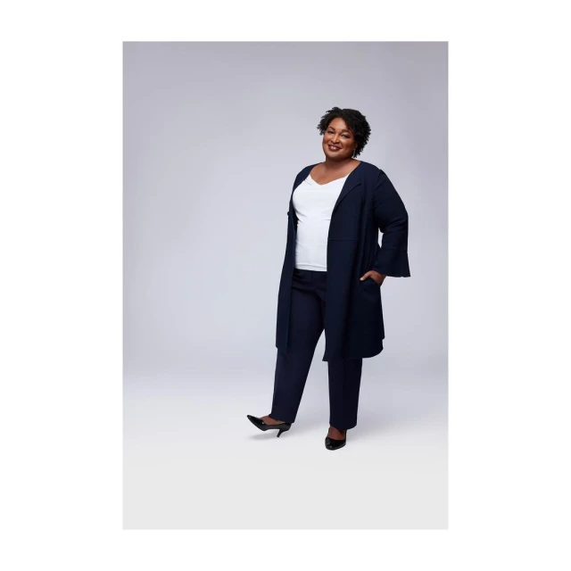Full-shot of a single adult woman in a dark-blue pant suit and dark shoes, standing and smiling in a photo studio against a grey background.