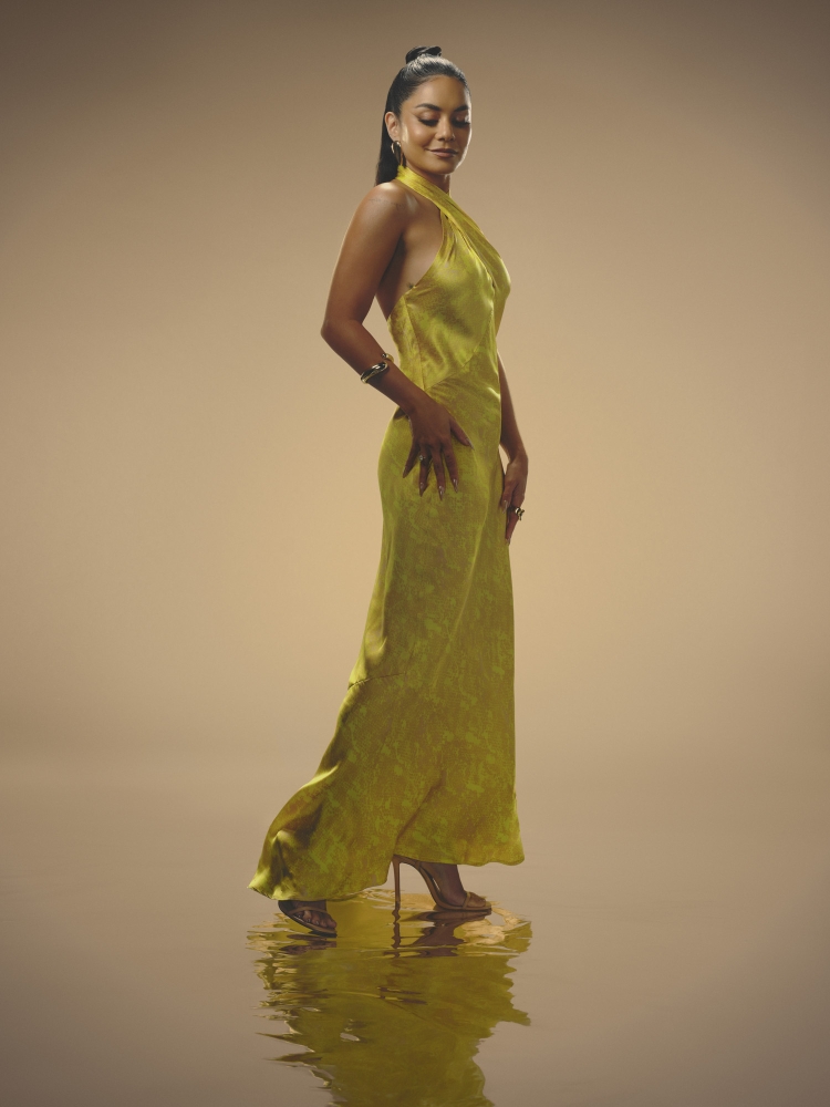 Elegant woman in a halter-neck chartreuse gown, stands in a photo studio, looking down with a reflective water-like surface beneath her.