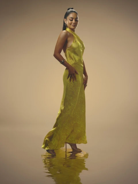 Elegant woman in a halter-neck chartreuse gown, stands in a photo studio, looking down with a reflective water-like surface beneath her.