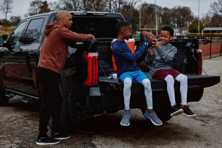 Two children sitting on a pickup truck tailgate playing with a football, with an adult male standing next to the truck outdoors.