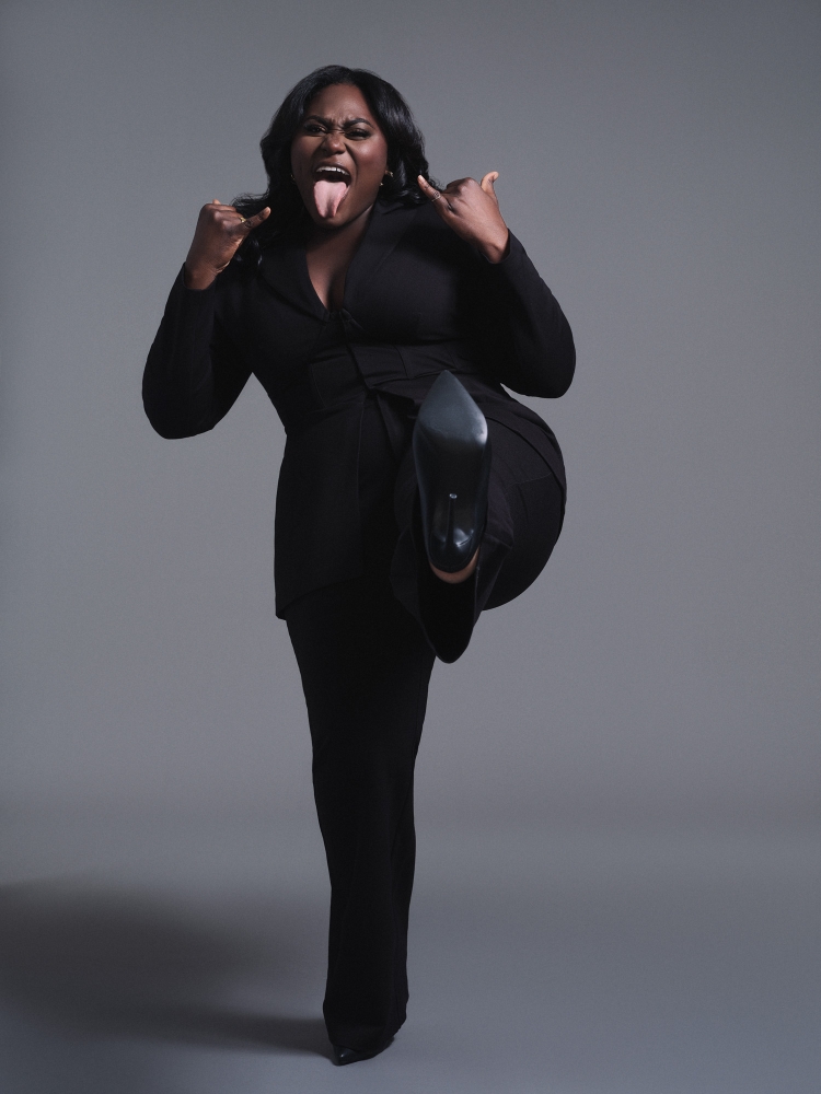 An adult female model wearing a black suit, showing her tongue and holding up a pointed-toe stiletto shoe against a grey background.