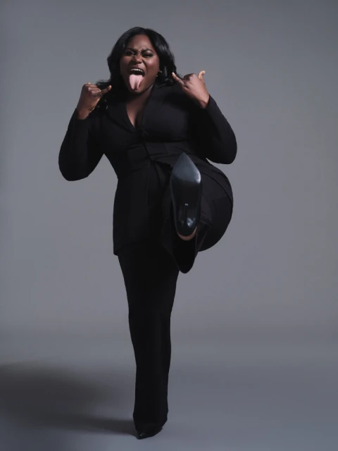 An adult female model wearing a black suit, showing her tongue and holding up a pointed-toe stiletto shoe against a grey background.