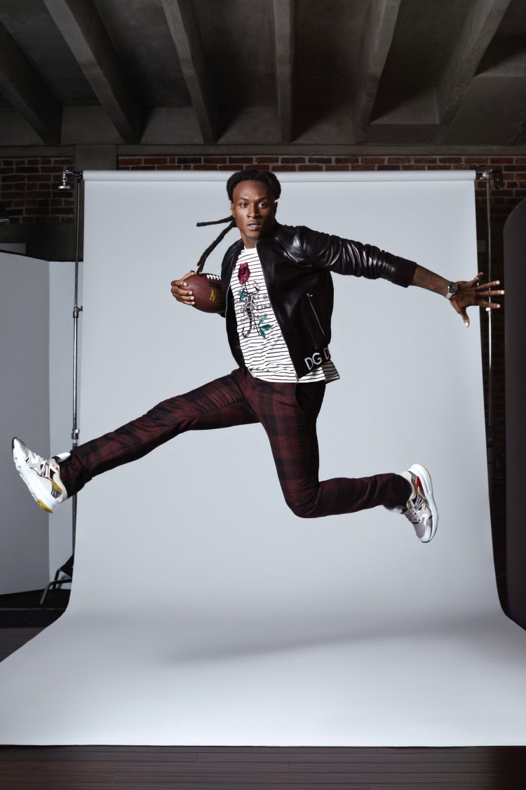 African American male in a black leather jacket and patterned pants leaps in a photo studio holding a football, showcasing dynamic energy.