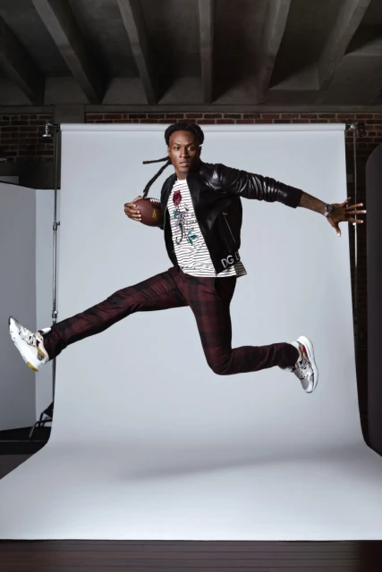 African American male in a black leather jacket and patterned pants leaps in a photo studio holding a football, showcasing dynamic energy.