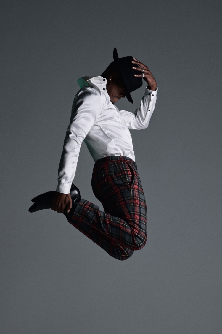 An African-American man in mid-air, captured in a dynamic studio shot, wearing a white shirt, plaid pants, black hat and black shoes.