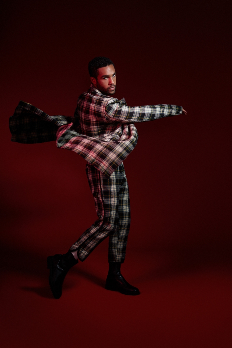A stylish Black male model in a plaid suit strikes a dynamic pose against a deep red backdrop, showcasing a unique fashion statement.