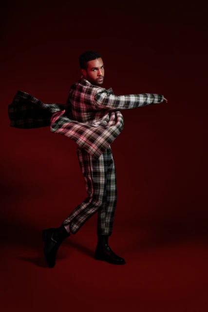 A stylish Black male model in a plaid suit strikes a dynamic pose against a deep red backdrop, showcasing a unique fashion statement.