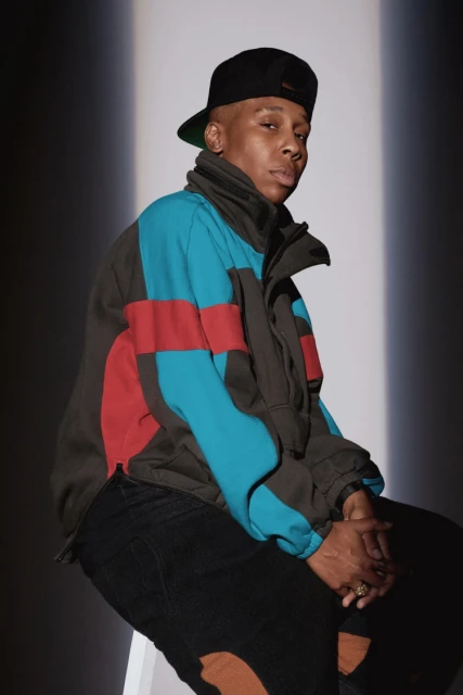 Portrait of a stylish adult male with a cap and color-blocked jacket sitting indoors with neutral lighting and a shadow backdrop.