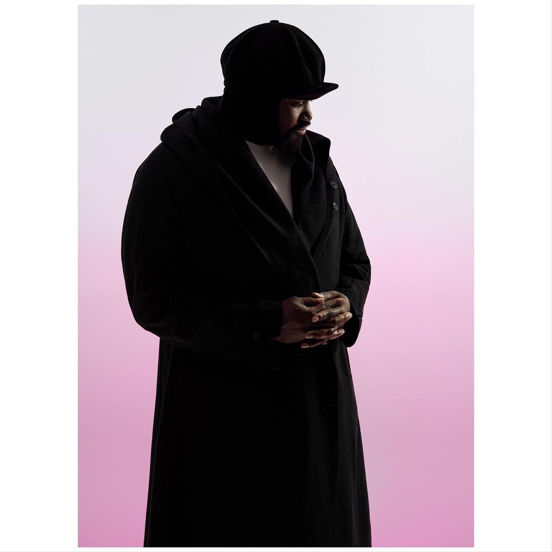 Portrait of an African American man wearing a black hat and long coat against a gradient background of white and pink.