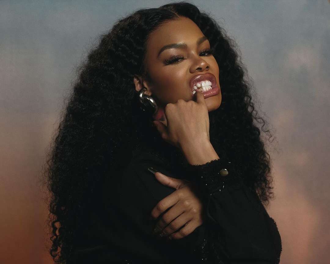Portrait of African-American female with long black curly hair and gold teeth, wearing black top and large earrings against blurred background.