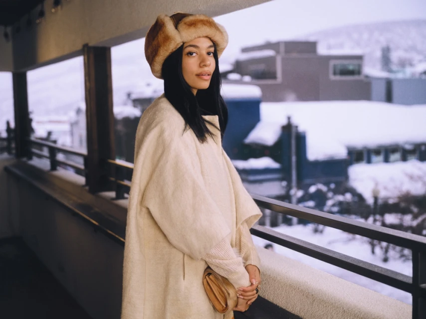Adult female in winter hat and cream coat stands on balcony with snow-covered mountain landscape in the background on a cloudy day.