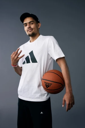 Basketball player Trae Young wearing a white t-shirt and black cap holding a basketball against a grey background