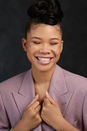 Close-up portrait of a smiling woman with an elaborate updo hairstyle wearing a mauve blazer against a dark textured background.